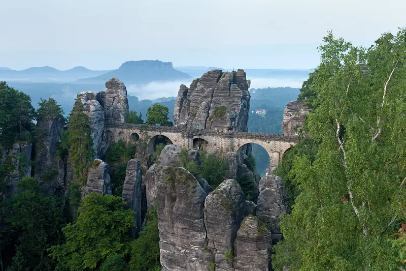 Die steinerne Basteibrücke zwischen Sandsteintürmen im Morgenlicht, dahinter Nebel im Tal